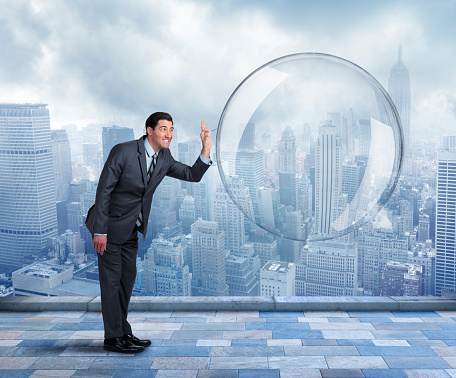 A businessman, holding a needle, is about to pop a large over sized bubble.  He stands in front of the New York City skyline on a cloudy and dreary day.