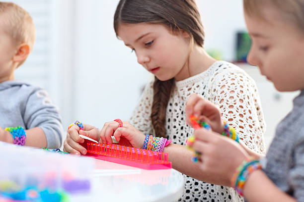 Little children sitting together at a table making bracelet from rubber loom bands at home