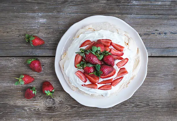 Rustic Pavlova cake with fresh strawberries and whipped cream over a rough wood background. Top view