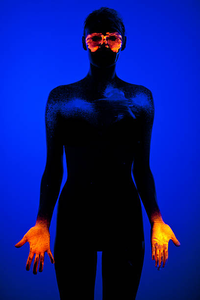 Creative portrait of young woman painted with fluorescent makeup that glowing under the ultraviolet light. Makeup contains orange mask  around the eyes, glowing hands and some discrete blue parts on body. Background is also blue, illuminated with blue neon light, but this light makes her looks like silhouette exept painted parts. Photo taken under the real neon ultraviolet light (black light). High ISO value