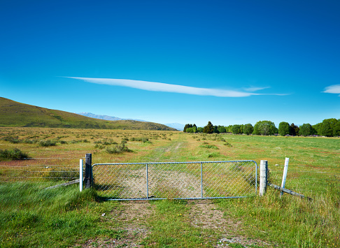 A gate blocks the way onto a farm track that stretches off into the distance. This scene of farmland is in Mackenzie Country, South Canterbury, on New Zealand's South Island. Off in the distance can be seen the snow-capped peaks of the Southern Alps. Composite image.