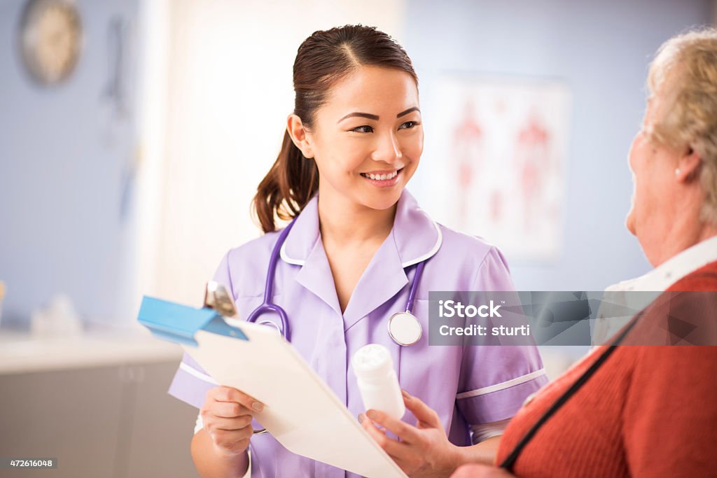 happy nurse chats to senior patient a young nurse stands and chats to a senior woman patient as she leaves the surgery. She is wearing a lilac nursing uniform and holding a bottle of pills . She is smiling to the senior woman and explaining . In the background a hospital interior can be seen. 20-29 Years Stock Photo