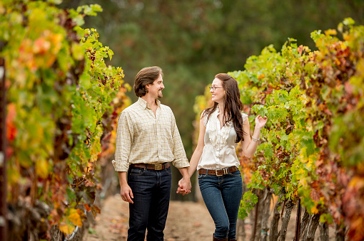 A heterosexual couple holding hands and walking together on a path looking at the grapes at a vineyard in the Napa Valley while on their honeymoon vacation.