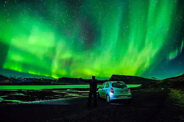 Nordic nightscape and a backlit human figure spreading arms near parked car under a sensational celestial lights eruption Aurora Borealis, which makes Iceland popular spot for tourist willing to witness one of the greatest natural phenomenon. Shot with Canon EOS system, wide angle lens, ISO, long exposure.