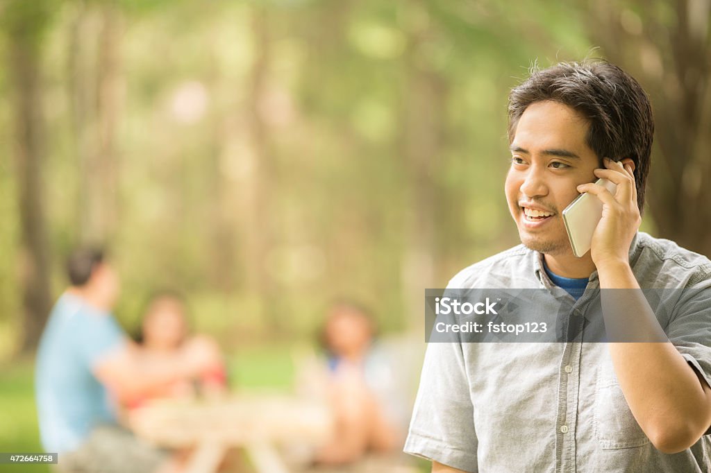 Happy young adult man talking on smart phone outdoors. Summer. Happy, laughing young adult man talking on his smart phone outdoors in the summer or spring season. The Asian descent man is all smiles as he chats with a friend using his mobile device. Group of unrecognizable people in the background sit at an outdoor table. Park or campus setting. Beautiful nature background.  20-29 Years Stock Photo