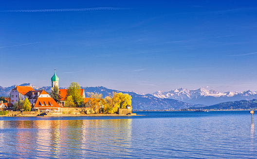 The picturesque peninsula village of Wasserburg with the landmark Church of St George and a passenger ferry in front of the majestic snow-covered Swiss alps at Lake Constance (Bodensee).
