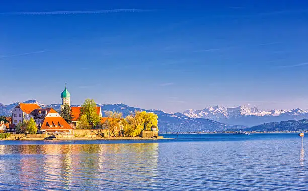 The picturesque peninsula village of Wasserburg with the landmark Church of St George and a passenger ferry in front of the majestic snow-covered Swiss alps at Lake Constance (Bodensee).