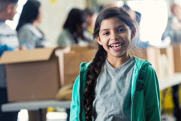 Cute little girl smiling in local food bank Elementary age Hispanic girl is smiling in front of assembly line in local food bank. She has a long braid and is wearing a green hooded sweatshirt. Diverse volunteers are packing cardboard boxes with donated groceries in background. word processing stock pictures, royalty-free photos & images