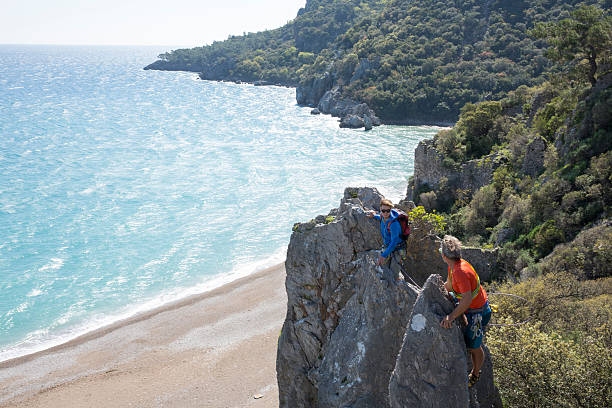 Two climbers stand on knife edge ridge above beach and sea, greeting each other