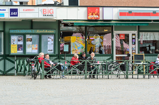 Karlshamn, Sweden - May 06, 2015: Senior persons sitting outside with gloomy faces and relaxing. Shops in background. Fenced in area around persons.