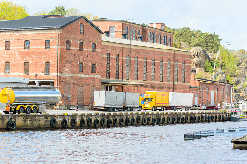 Karlshamn, Sweden - May 06, 2015: Trucks parked outside historic brick factory building waiting to unload their cargo. Unknown worker walking outside. Water in foreground.