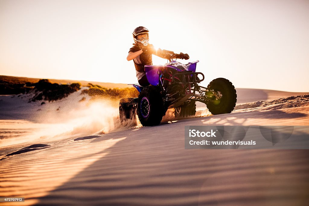 Quad racer driving up a sand dune with sun flare Competitive quad bike racer kicking up sand while driving up a sand dune on a summer evening with sun flare Quadbike Stock Photo