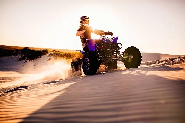 Competitive quad bike racer kicking up sand while driving up a sand dune on a summer evening with sun flare