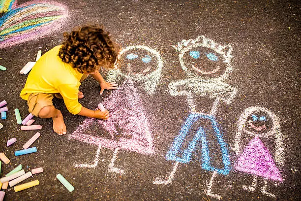 Overhead shot of a little boy drawing a  picture of his family in colourful chalk on tarmac
