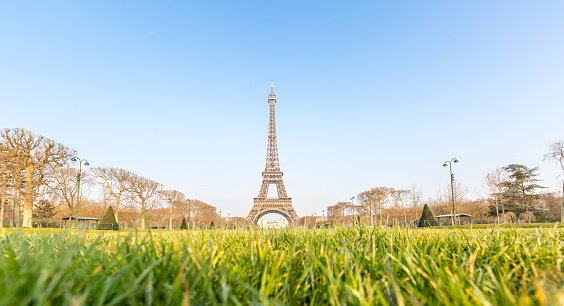 A digital photograph of the Eiffel Tower structure in Paris, France, as viewed from the Champ-de-Mars in perspective with clear blue, cloudless, skies serving as background during a vibrant sunny afternoon. Few people in the photo allowing for a clearer view of the grass and fading treeline that give in to the tower.