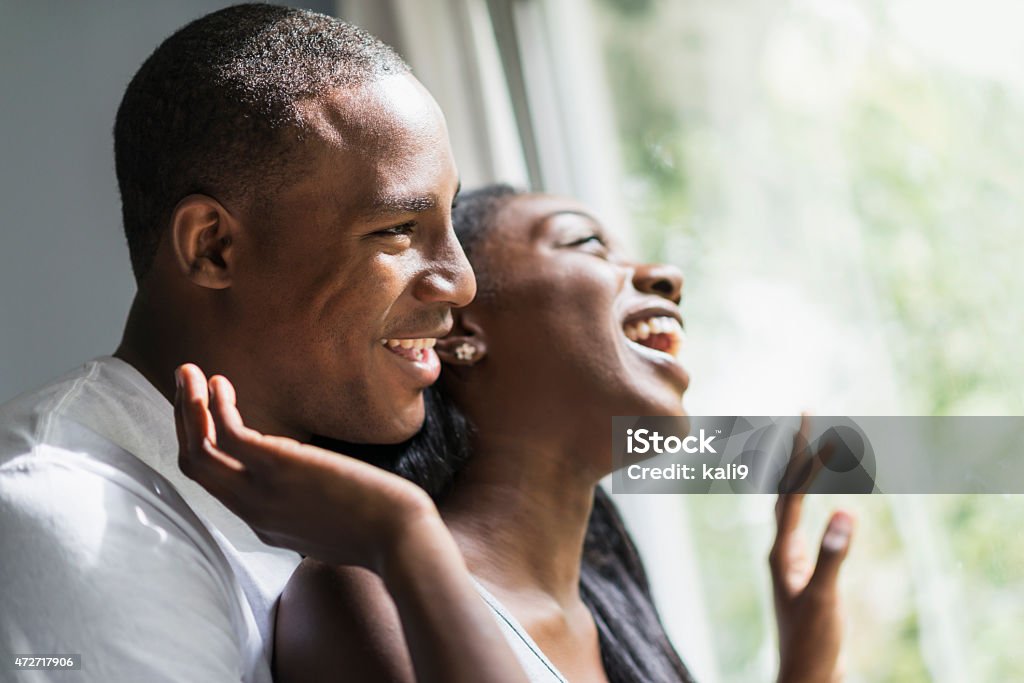 Faces of young black couple looking out window, laughing Close up of a young, happy African American couple.  The man is standing behind the woman and they are laughing as they look out the sunny window.  She has one hand on the glass and is reaching behind her to touch her boyfriend's face. Close To Stock Photo