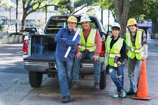 A group of four diverse workers wearing hard hats and yellow safety vests with pickup truck on city street.  They are a crew of construction or utility workers, smiling at the camera.  One of the workers is a woman and two are Hispanic.