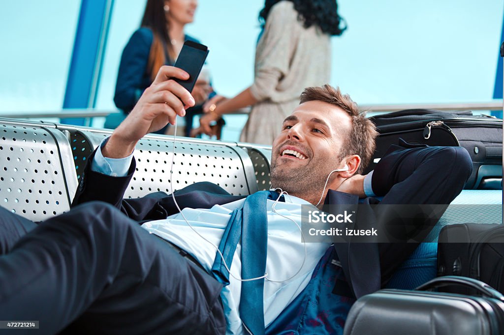 Businessman waiting for the flight, using smart phone Happy businessman wearing suit and earphone lying down on bench at he airport lounge, holding smart phone in hand, suitcase next to him. People in the background. 2015 Stock Photo
