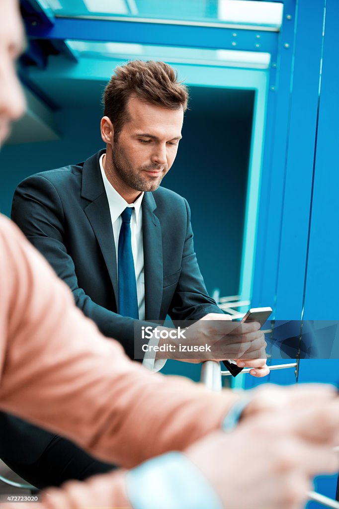 Businessman texting on smart phone in an office hall An attractive businessman in a suit texting on smart phone in an office with blured man in the foreground. 2015 Stock Photo
