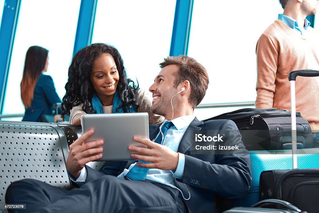 Businessman waiting for the flight, watching movie on digital tablet People waiting for a flight at the airport lounge. On the foreground smiling businessman wearing suit and earphone lying down on bench, holding digital tablet in hand and talking with arfo amercian smiling woman. 2015 Stock Photo