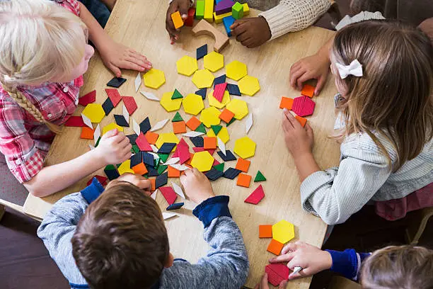 An overhead view of a group of five preschoolers sitting at a table playing with colorful blocks and geometric shapes.  They are unrecognizable since we see the tops of their heads and their hands.  They are in a preschool or kindergarden classroom.