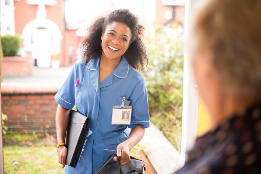 A female nurse or community care worker is at the front door of her senior female patient and saying hello. She is wearing a blue nurses tunic , and holding a medicines bag . She is wearing an ID badge with her profile photo already on it . The senior patient has her back to us at the front door . In the background a residential street can be seen defocussed .
