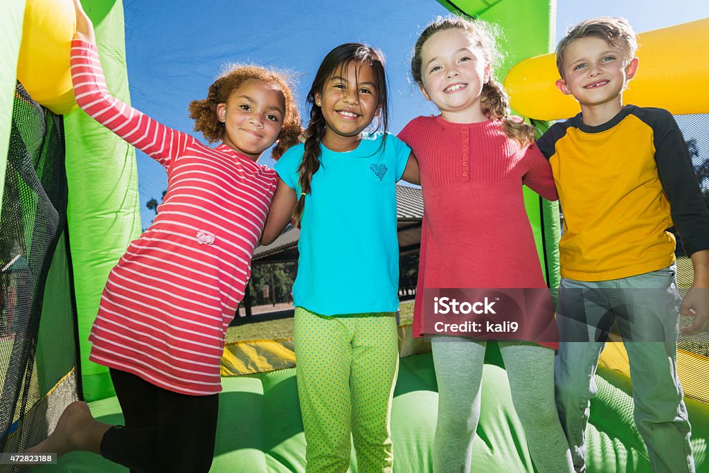 Four multi-ethnic children playing on bouncy castle A group of four multiracial friends having fun on a bounce house.  They are standing side by side with their arms around each other's shoulders, smiling at the camera.  There is one boy and three girls, 6 to 8 years old.  The inflatable castle is bright yellow and green. Bouncy Castle Stock Photo