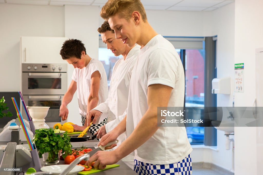 Group of Student Chefs Learning with Instructor A horizontal image of 2 young chef students dressed in chef uniforms and aprons. There is an older male teaching the students and demonstrating what to do. Cooking Stock Photo