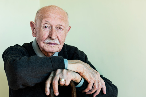 Candid portrait of a sprightly looking 90 year old man. Horizontal portrait with the subject placed off-centre giving some copy space to the left. He is looking to camera with a content expression on his face. He is wearing a green jumper over a shirt and tie, he has grey thinning hair and a white moustache, very shallow focus on his eyes.