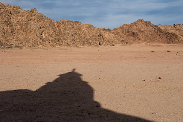 Shadow of a minaret of a small bedouin mosque in the egyptian desert. Sand and stones, rocky mountains in the background. Blue sky with clouds.