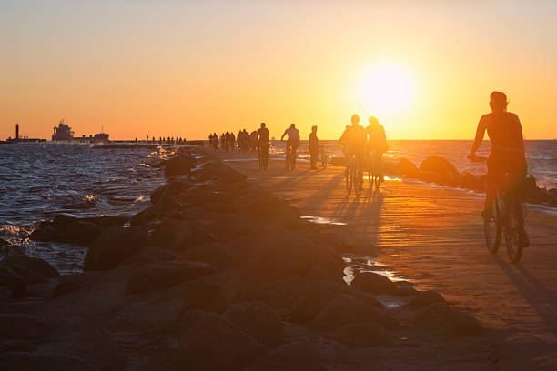People on bikes ride across the causeway to the lighthouse in the Baltic Sea with ships at sunset