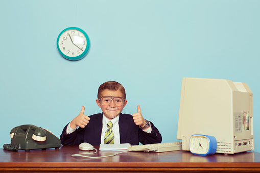 A young boy dressed as a businessman gives the thumbs up sign as there is some good business happening. He sits at his desk with retro computer and phone.