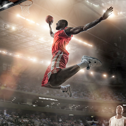 A low angle view of a professional basketball player jumping in mid air with arms outstretched and basketball held high ready to slam dunk whilst being looked at by rival player. The action occurs in a basketball game in a generic floodlit indoor basketball arena full of spectators. With motion blur and lensflare. 
