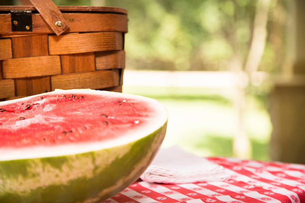 Sliced watermelon outdoors on picnic table. Summer. Basket. A red, ripe slice of watermelon on picnic table outdoors in summer season. A picnic basket is behind the watermelon, which lays on a red checked tablecloth. Beautiful and sunny park, nature scene in the background.  No people in image.  Memorial Day, July 4th, or Labor Day family picnic concept. Copyspace to right.  june stock pictures, royalty-free photos & images