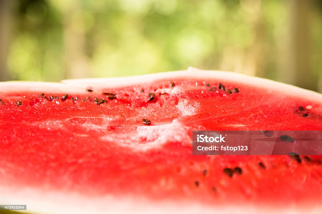 Close-up sliced watermelon outdoors on picnic table. Summer. A close-up view of a red, ripe slice of watermelon on a picnic table outdoors in summer season. The watermelon is juicy with seeds. Beautiful and sunny park, nature scene in the background.  No people in image.  Memorial Day, July 4th, or Labor Day family picnic concept.   2015 Stock Photo