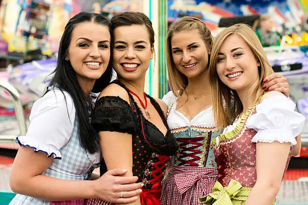 Joyful young and attractive women at German funfair Beer Fest with traditional dirndl dresses and joyride in the background. Mixed nationalities, 2 girls rather typical German, one with Asian and Caucasian blend and one Russian girl, could also be Greek.