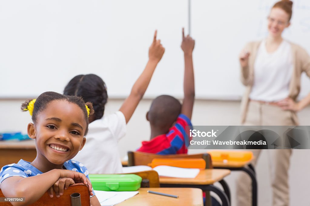 A little girl sitting in a classroom with a teacher Cute pupil smiling at camera in classroom at the elementary school Discussion Stock Photo