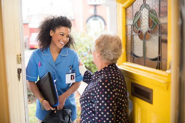 A female nurse or community care worker is at the front door of her senior female patient and saying hello. She is wearing a blue nurses tunic , and holding a medicines bag . She is wearing an ID badge with her profile photo already on it . The senior patient has her back to us at the front door . In the background a residential street can be seen defocussed .