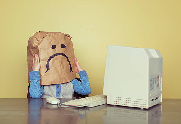 Sad Paper Bag Boy is Cyber Bullying Victim A young boy sits at a retro computer with a paper bag over his head. The paper bag has a large frown face as he has been a victim of online bullying. ugly face stock pictures, royalty-free photos & images