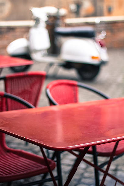 Red cafe tables and chairs on black cobbles, with an antique white scooter in the blurred-out background. Soft selective focus with copy space on the red table in the foreground. Shot in Rome, Italy