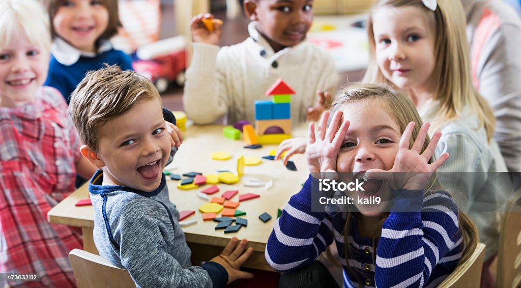 Playful preschoolers having fun making faces A multiracial group of preschoolers or kindergarteners having fun in the classroom.  Six children are sitting around a little wooden table playing with colorful wooden block and geometric shapes.  The playful little girl in the foreground is making a silly face at the camera. Child Stock Photo
