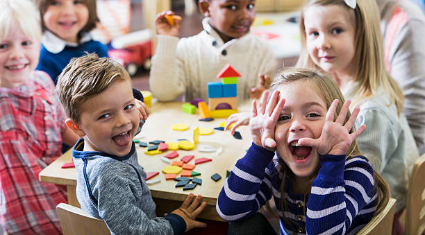Playful preschoolers having fun making faces A multiracial group of preschoolers or kindergarteners having fun in the classroom.  Six children are sitting around a little wooden table playing with colorful wooden block and geometric shapes.  The playful little girl in the foreground is making a silly face at the camera. preschool photos stock pictures, royalty-free photos & images