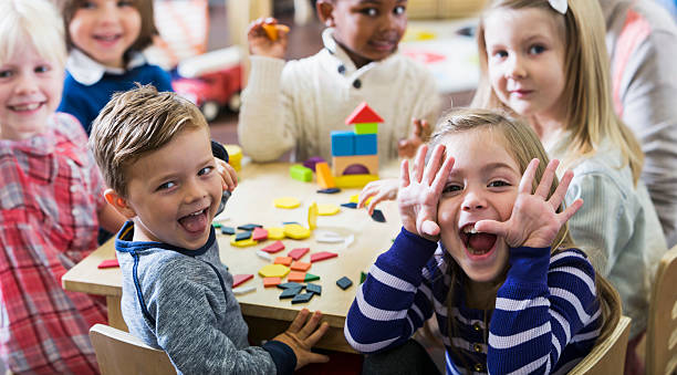 Playful preschoolers having fun making faces A multiracial group of preschoolers or kindergarteners having fun in the classroom.  Six children are sitting around a little wooden table playing with colorful wooden block and geometric shapes.  The playful little girl in the foreground is making a silly face at the camera. messing about stock pictures, royalty-free photos & images