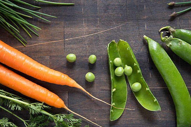 close up from above on arrangement of orange carrots , green snow pea  with open fruit and single peas that are spread on the table and green chive on dark rustic wooden background