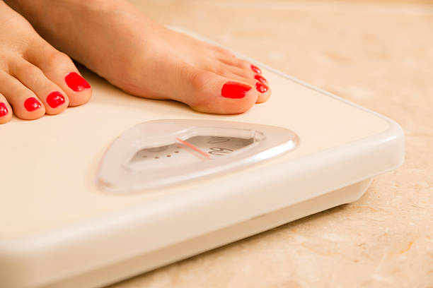 Close up of a caucasian woman with red painted toenails standing on a bathroom weight scale.  She is excited to read the scale, which shows that she has lost weight.  Dieting, weight loss concept.  Unrecognizable person. 