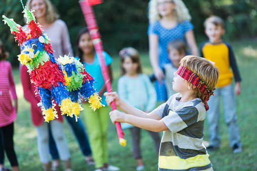A little boy, 6 years old, tries to hit a colorful pinata with a red stick at a birthday party or cinco de mayo celebration.  He is blindfolded.  A group of mothers and children watch in the background.