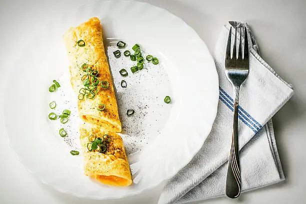 Breakfast. Rolled up, French style, egg Omlette served with fresh Scallion (green onion) and pepper on white plate. Ready to eat with fork and kitchen cloth. High angle view.