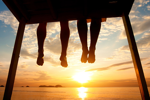 silhouette of feet of couple sitting on the pier at sunset beach, low angle view