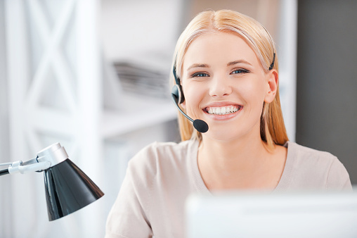 Happy young woman in headset looking at camera and smiling while sitting at her working place