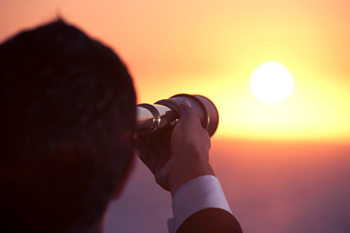 Businessman looking to the future with telescope at sunrise. He is holding the telescope to his eye and looking to the horizon over the ocean. This is a forecasting, discovery, visionary concept. It could also portray surveillance, opportunity, exploration and innovation.